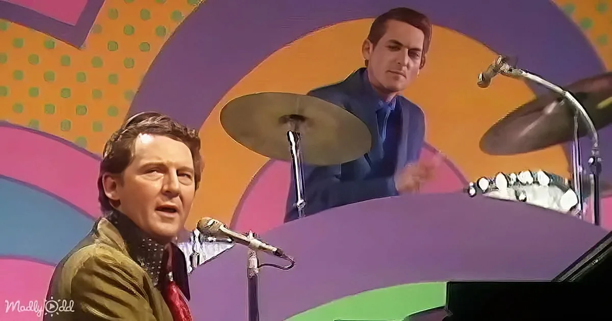 A man sings into a microphone on stage, with a colorful background and another person behind him.