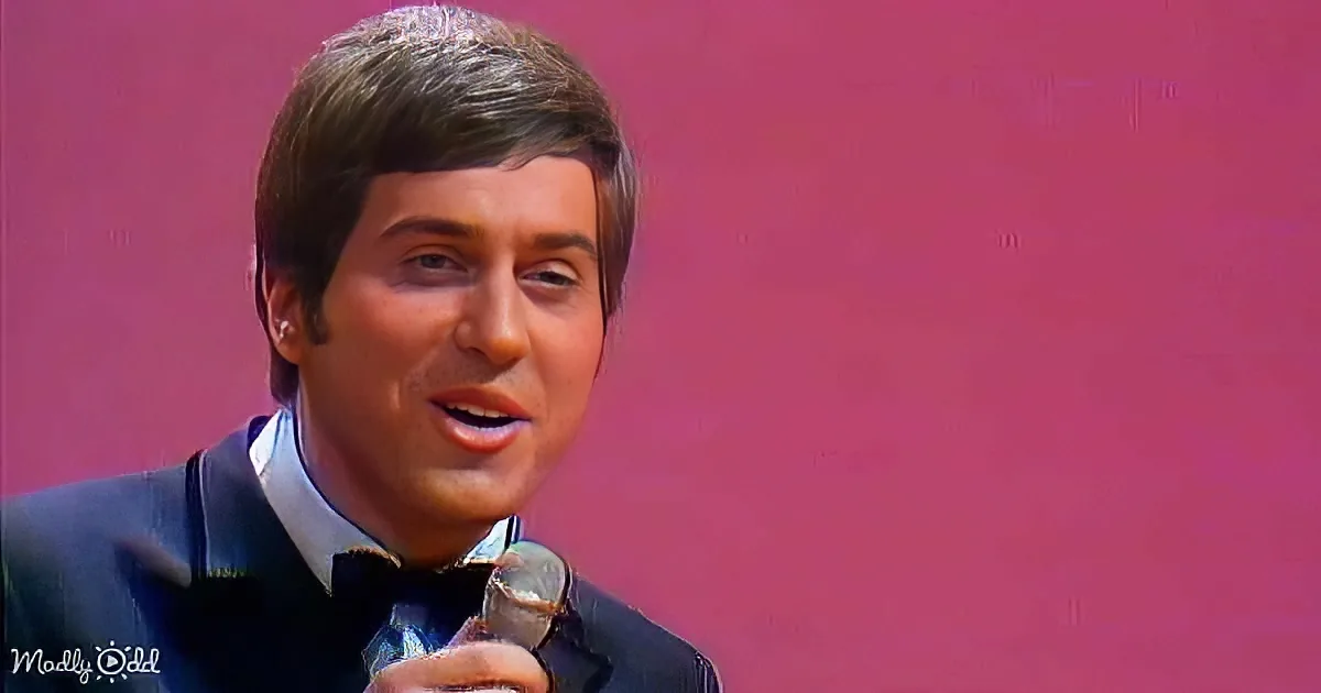 A man in a suit and bow tie holds a microphone, set against a pink background.