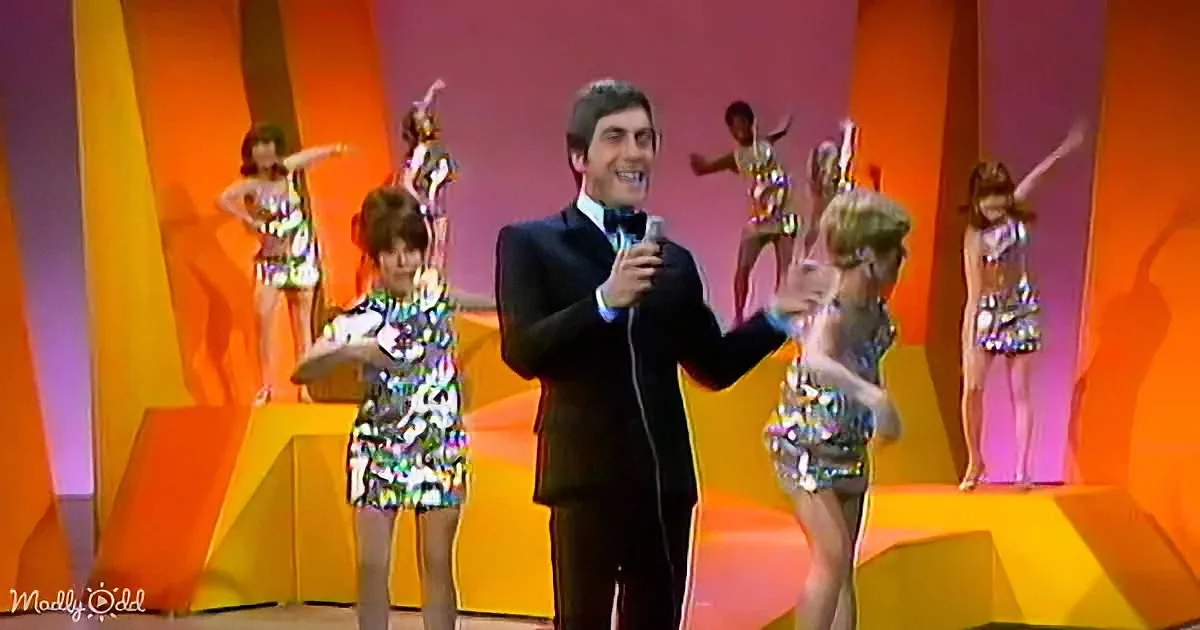 A man in a suit holds a microphone, smiling, with dancers in colorful outfits behind him on a vibrant stage.