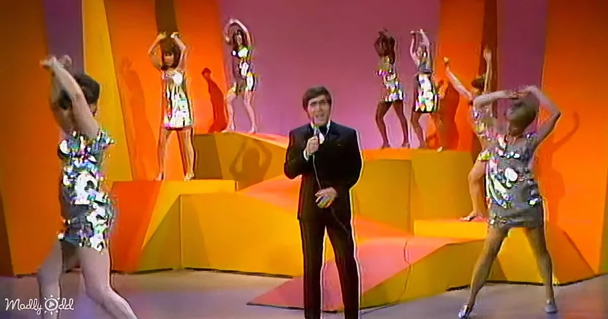 A man in a suit holds a microphone on stage, surrounded by dancers in colorful outfits.