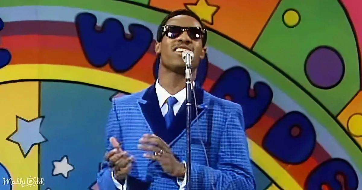 A man in a blue suit and sunglasses sings into a microphone against a colorful, star-patterned backdrop.