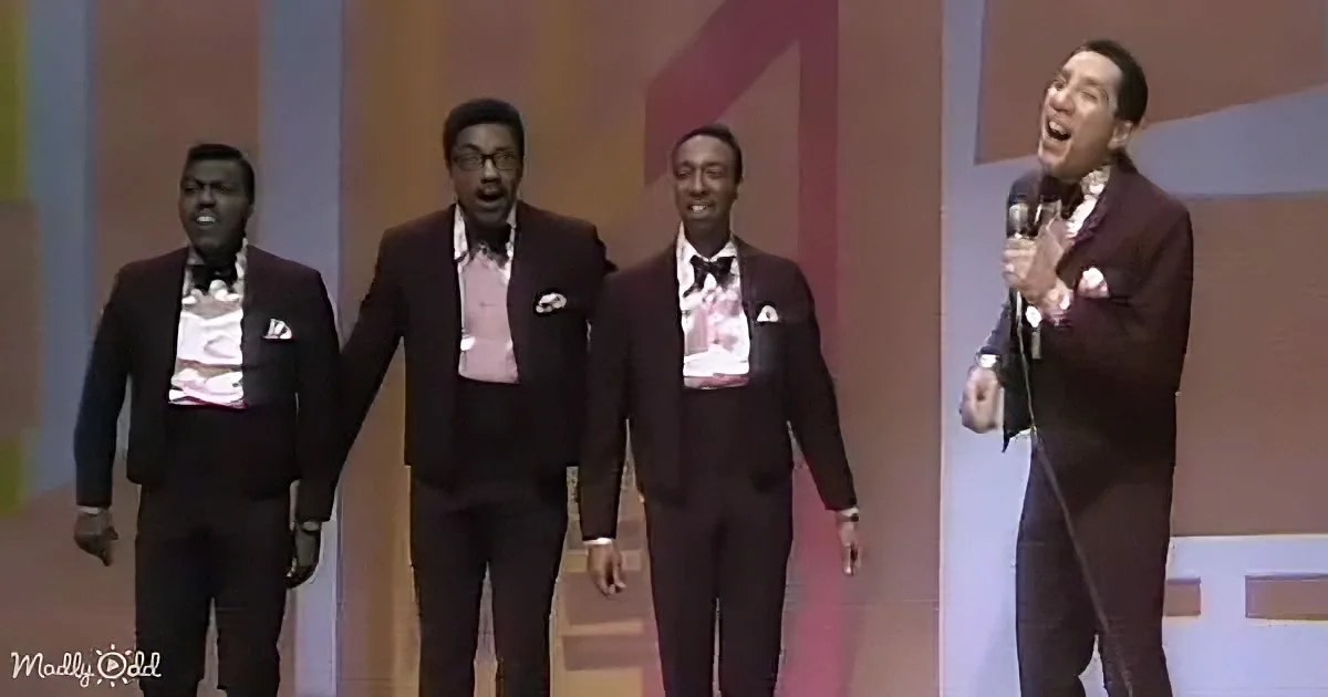 A man in a suit sings into a microphone, accompanied by three other men in suits on stage.
