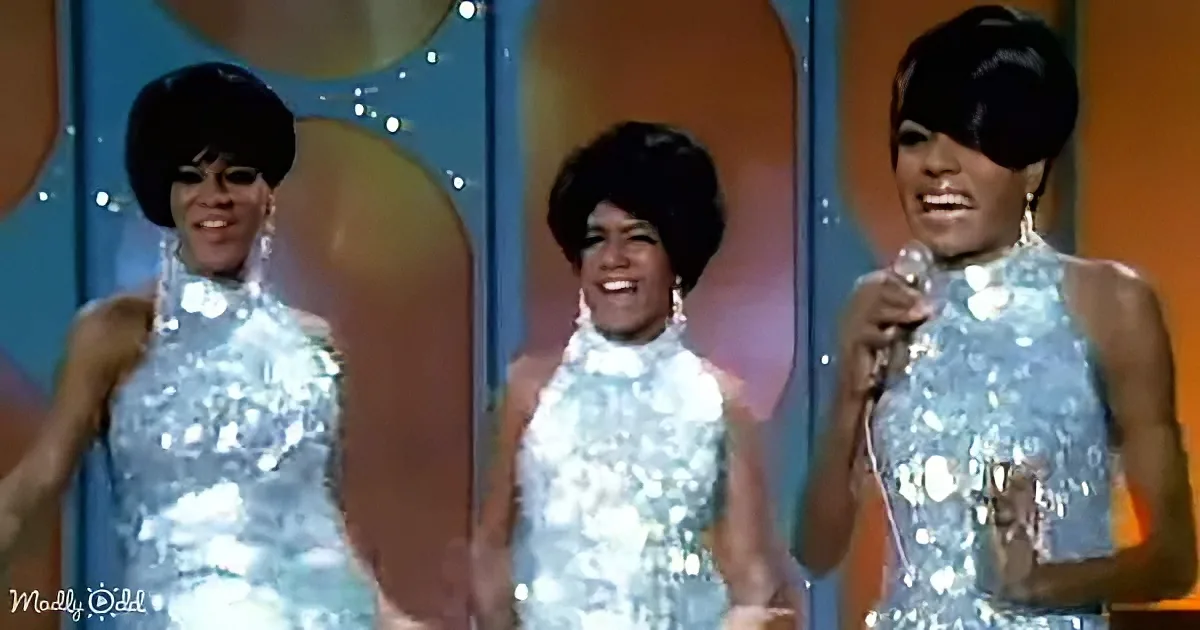 Three women in shiny silver dresses perform on stage, one holding a microphone.