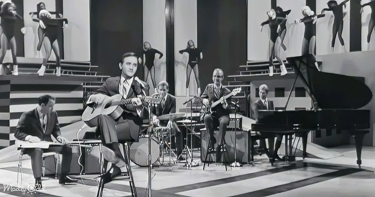 A man in a suit plays guitar on stage with a band and dancers in the background.