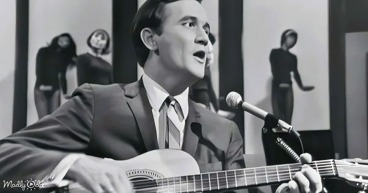 A man in a suit sings into a microphone while playing guitar, with dancers in the background.