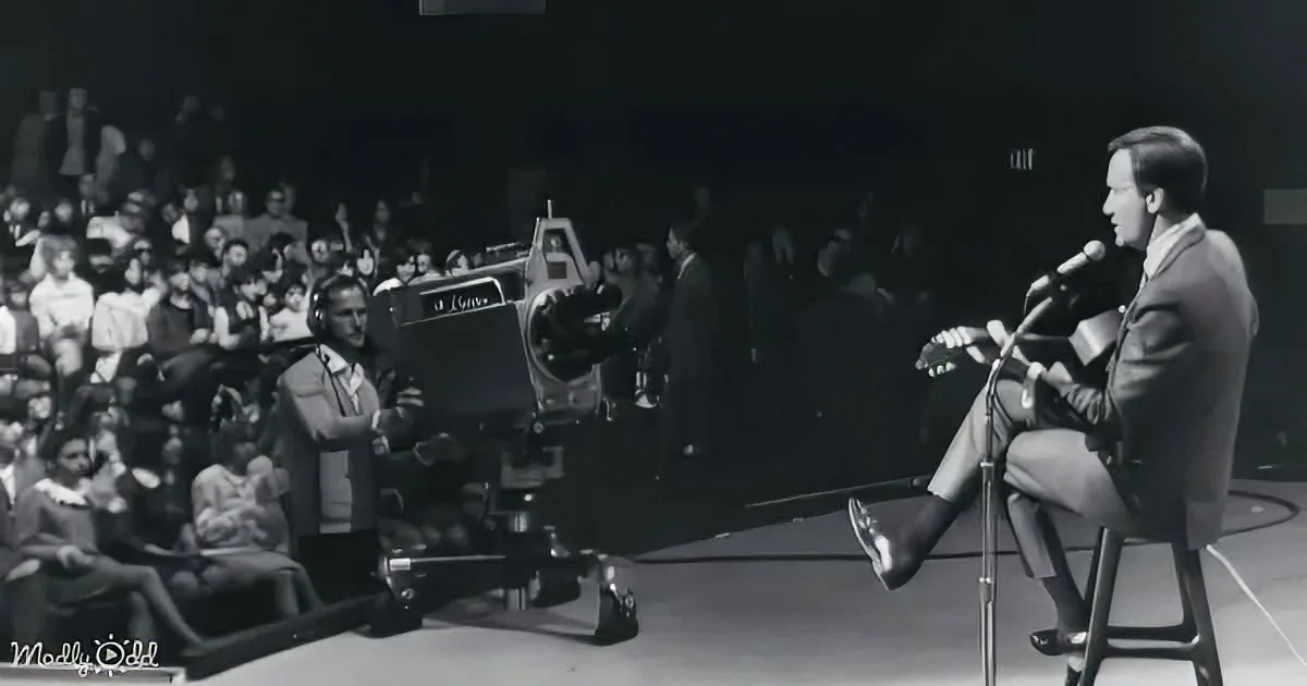 A man in a suit sits on stage with a guitar, addressing an audience, while a film crew captures the moment.
