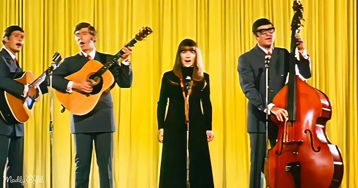 A band performs on stage with a yellow curtain backdrop, featuring a woman in a black dress and three men in suits, one...