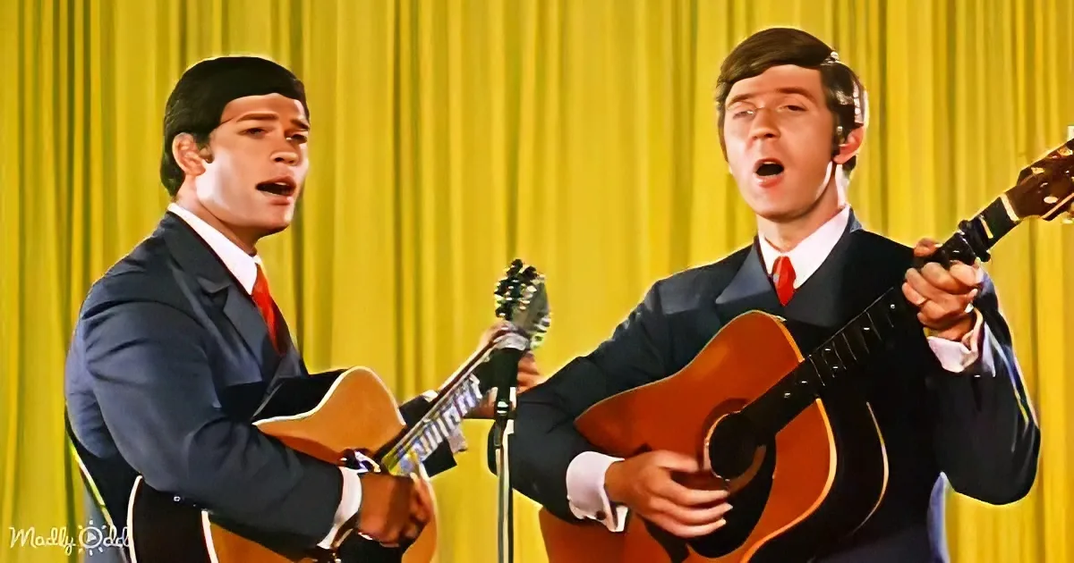 Two men in suits play guitars and sing on stage with a yellow curtain backdrop.