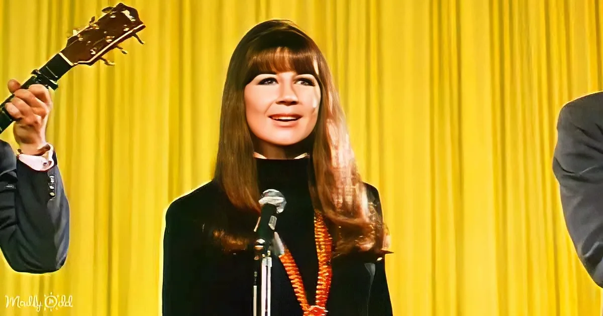 A woman with long brown hair, wearing a black outfit and an orange necklace, stands in front of a microphone with a...