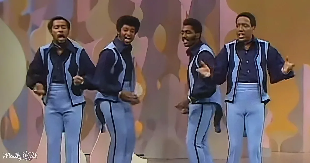 Four men in matching blue and black outfits perform on stage with colorful abstract background.
