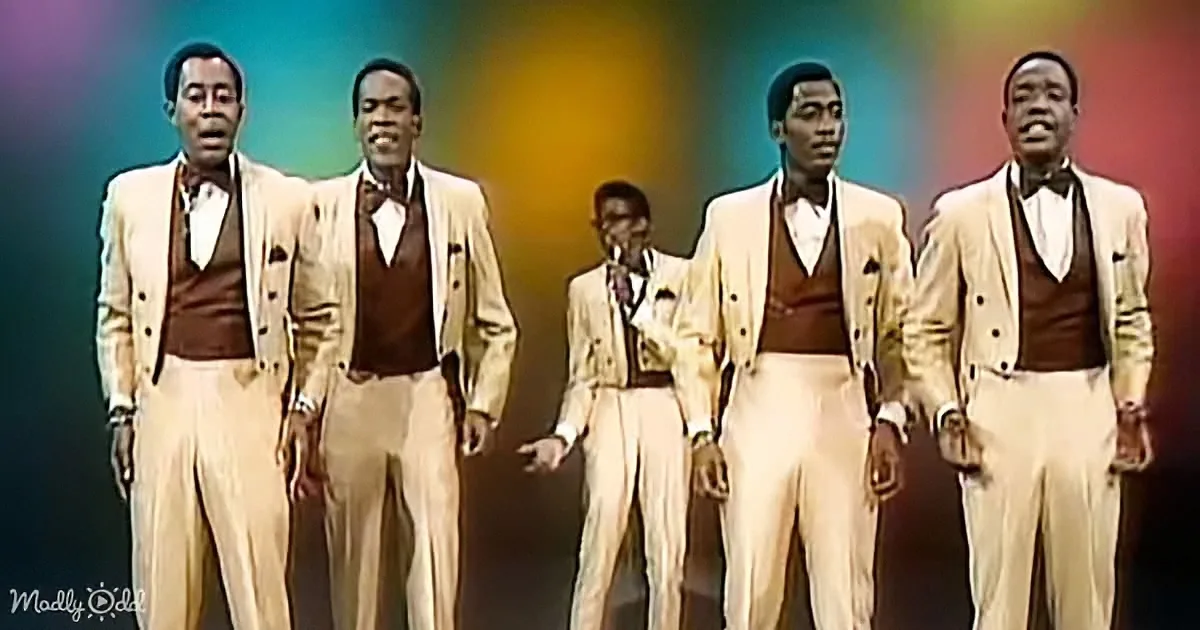 A group of five men in matching cream suits and brown vests stand in a line, performing on stage.