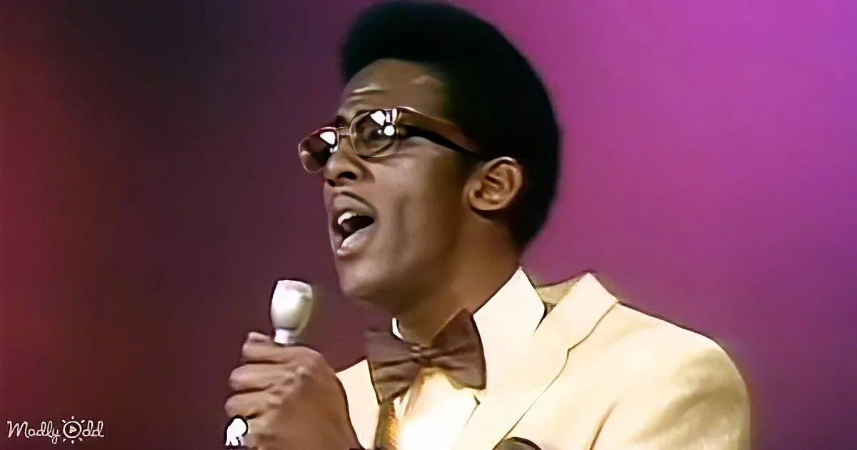 A man in a white suit and brown bow tie sings into a microphone against a purple background.