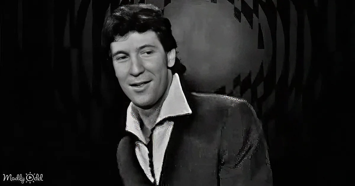 A man in a suit and open-collared shirt smiles, set against a dark, patterned background.