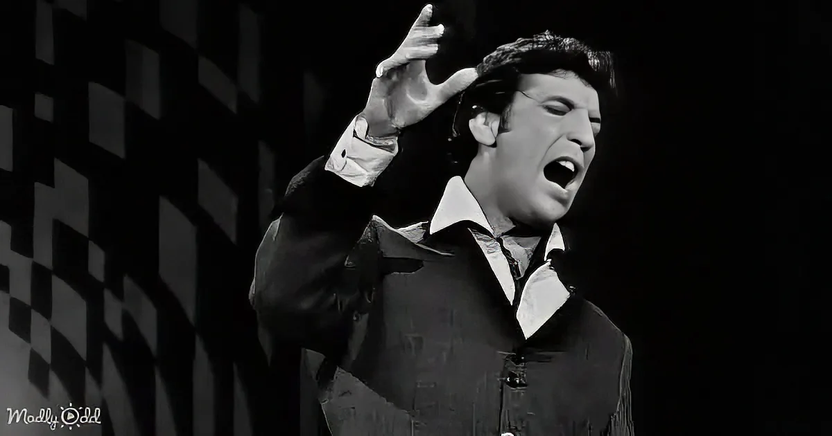 A man in a suit raises his arm, mouth open, against a checkered backdrop.