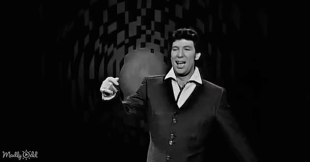 A man in a suit and tie is pointing and smiling, with a checkered background.