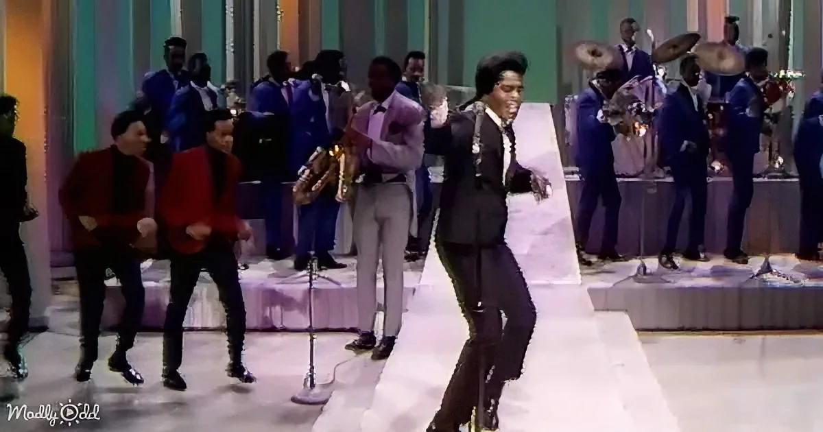A man in a black suit dances energetically on stage with a band in the background.