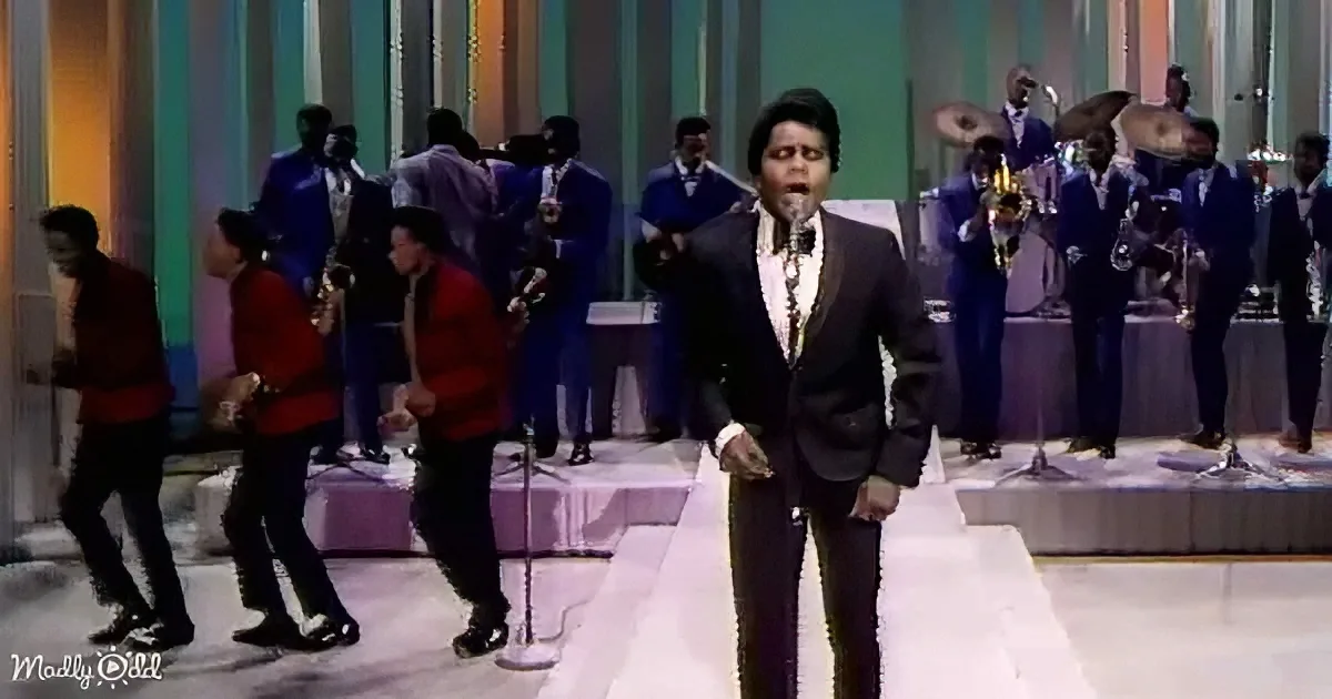 A man in a suit sings into a microphone on stage with a band and dancers behind him.