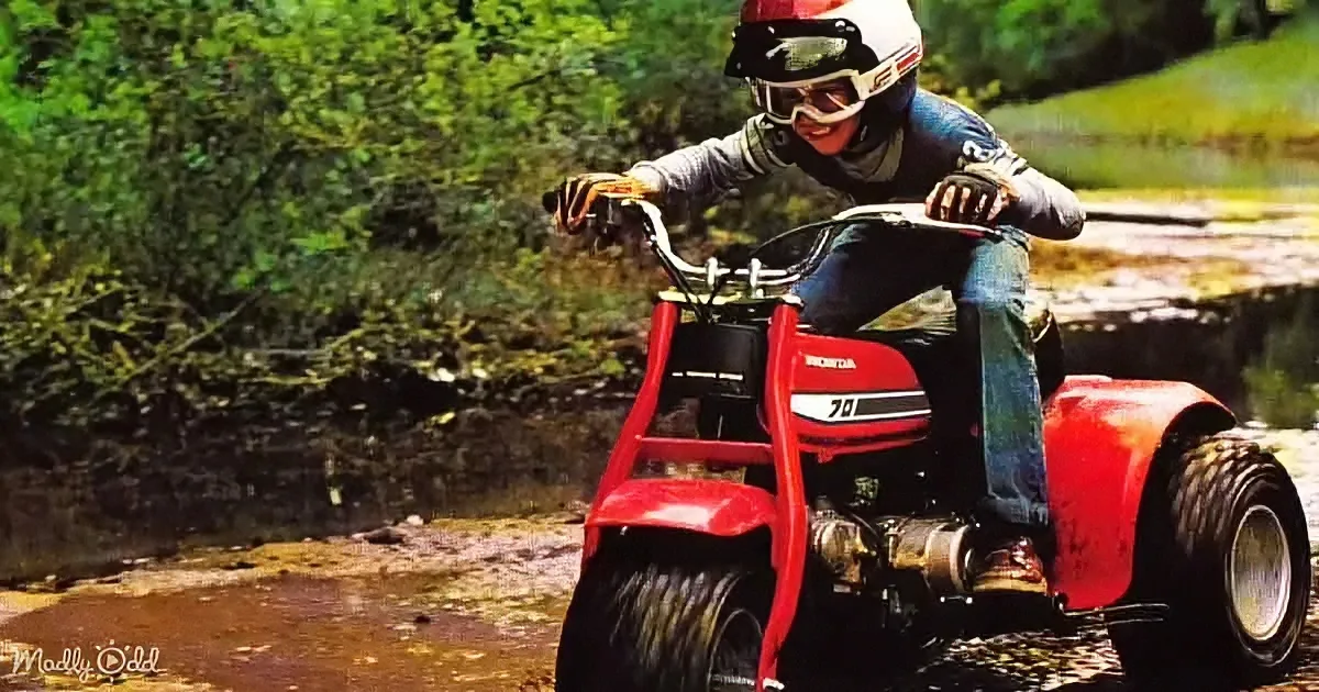 A person wearing a helmet and goggles rides a red Honda 700 ATV through a muddy stream.