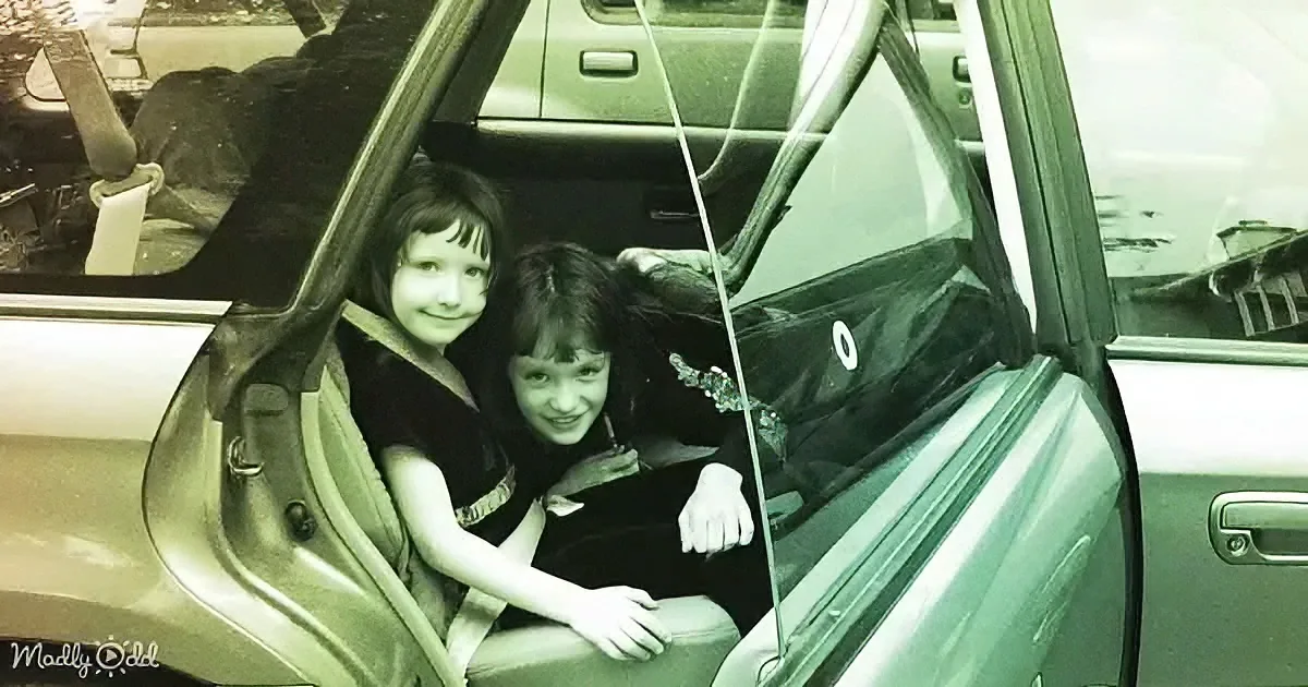 Two children smile from the backseat of a car with a seatbelt, looking out the open window.