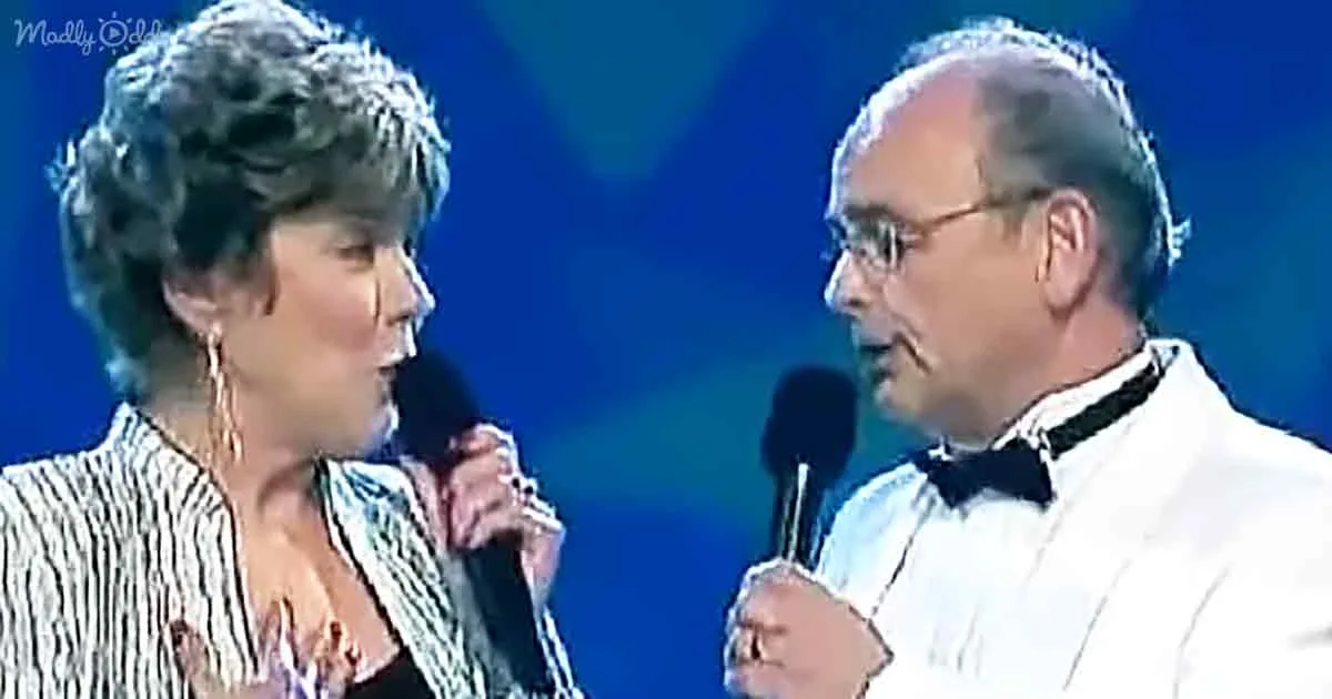 A man and woman, both holding microphones, appear to be singing or speaking on stage.