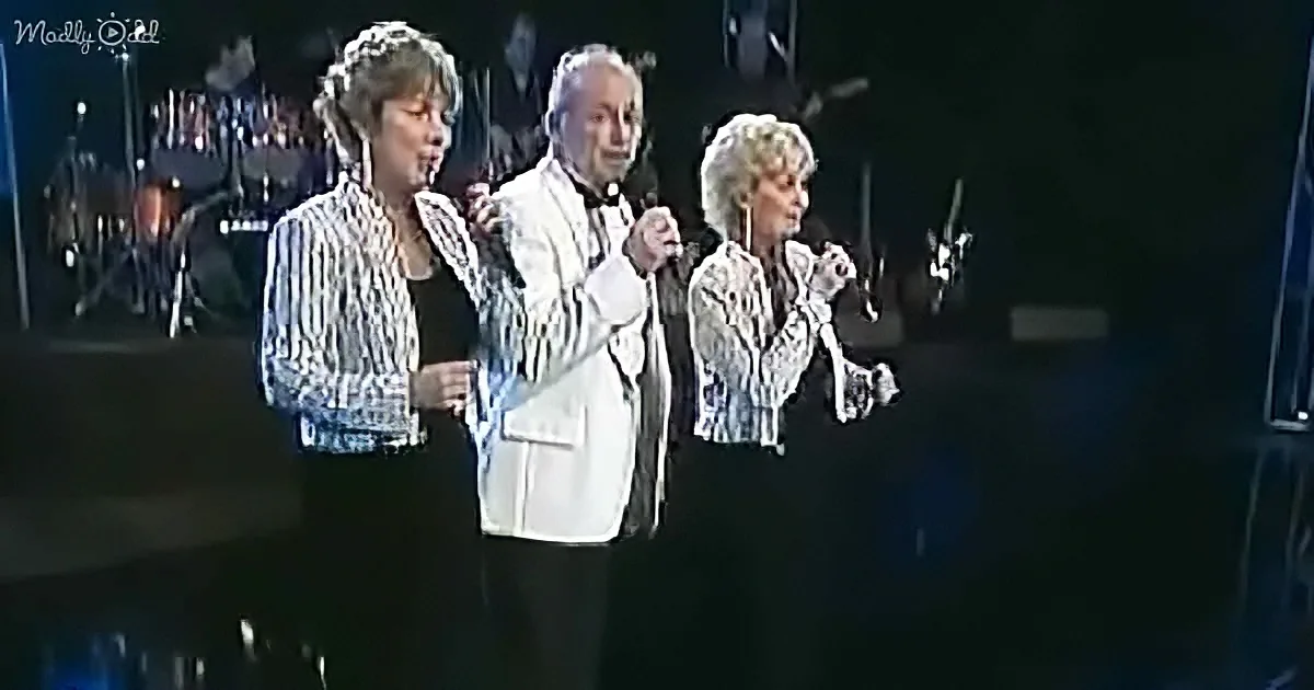 Three individuals in formal attire sing on stage, holding microphones, with a band in the background.