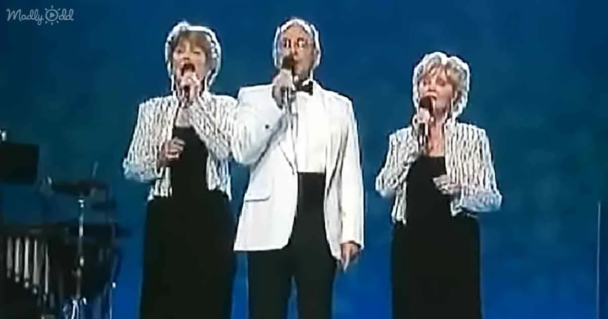 Three performers in formal attire sing on stage with microphones, illuminated by blue lighting.