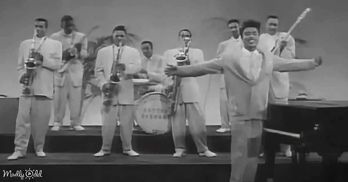 A man in a white suit sings passionately on stage with a band behind him.