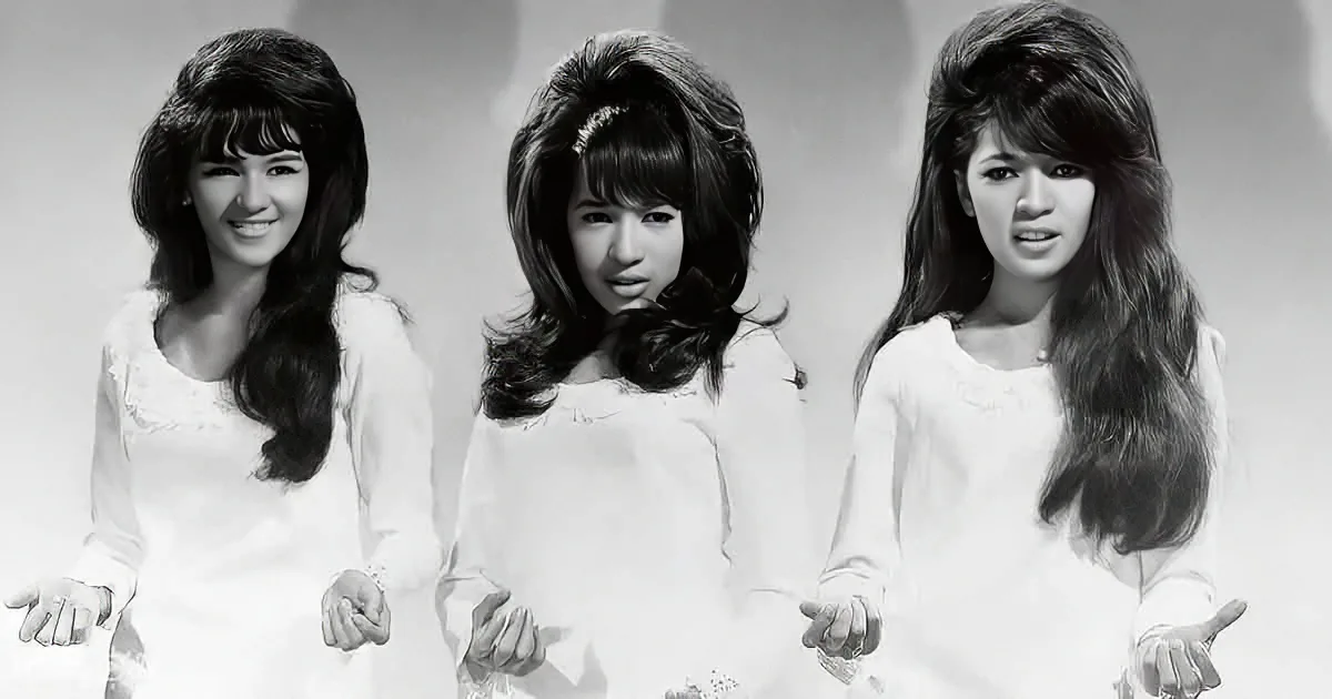 Three women in white dresses with long, wavy hair stand side by side, hands outstretched.