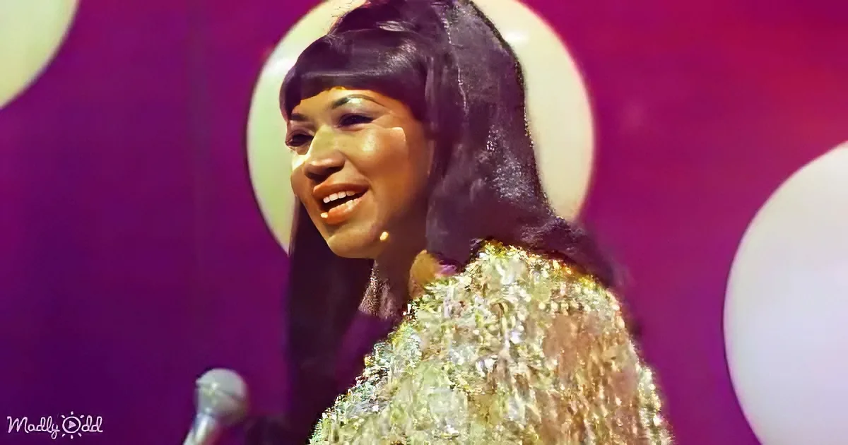 A woman in a glittery dress smiles on stage with a microphone and purple background.