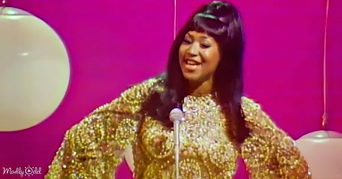 A woman in a glittery gold dress stands at a microphone, smiling, with a pink backdrop and a white balloon.