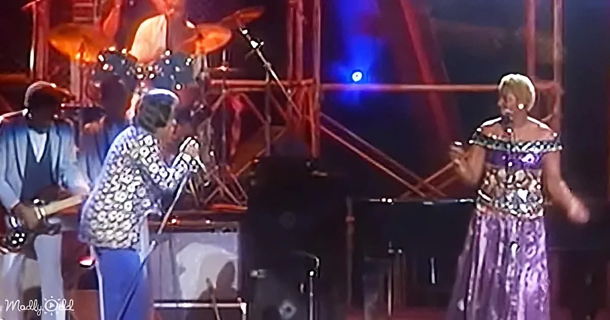 A woman in a sparkly dress sings on stage, accompanied by musicians and a drum set.