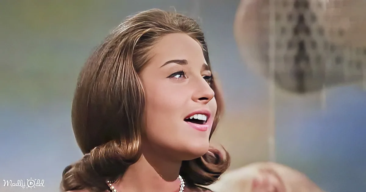 A woman with brown hair styled in a 1960s fashion, wearing a pearl necklace, looks upwards with a slight smile.