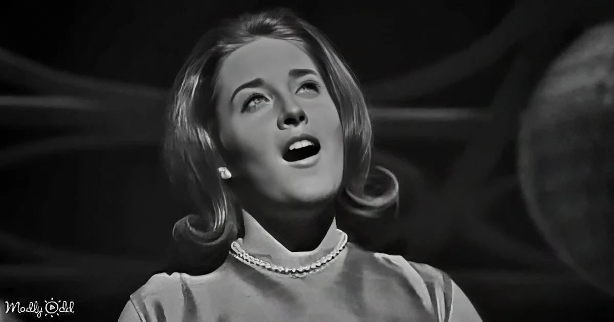 A woman with short, wavy hair and a pearl necklace is looking upwards with her mouth slightly open.