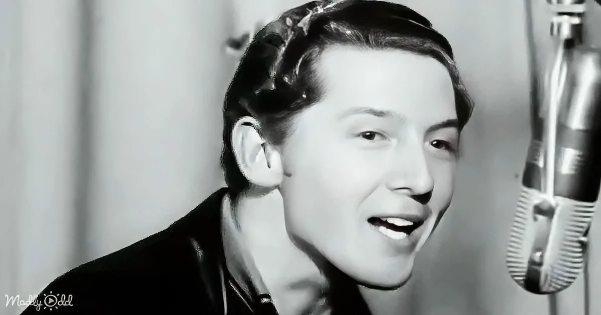 A man with slicked-back hair sings into a microphone, wearing a dark jacket, in a black-and-white image.