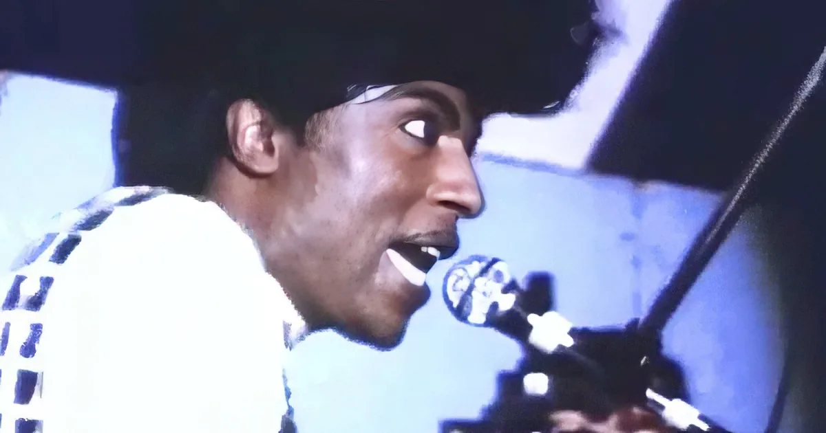 A person with an afro sings into a microphone, wearing a white shirt with black patterns.