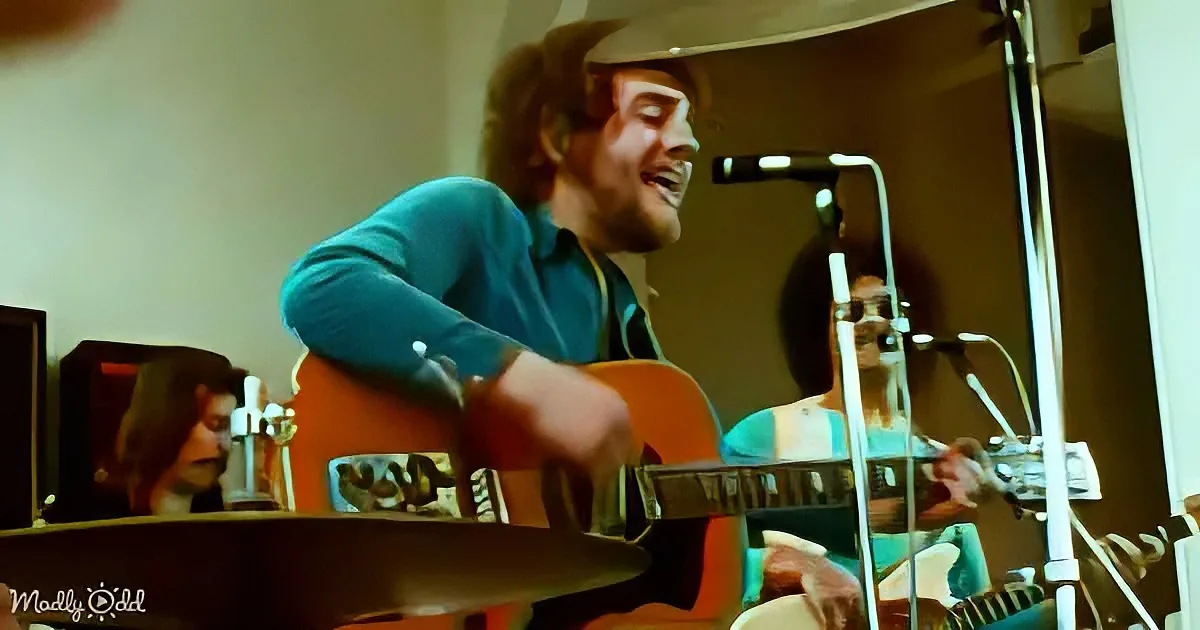 A man in a blue shirt and cowboy hat plays guitar in a recording studio with a microphone and other equipment.