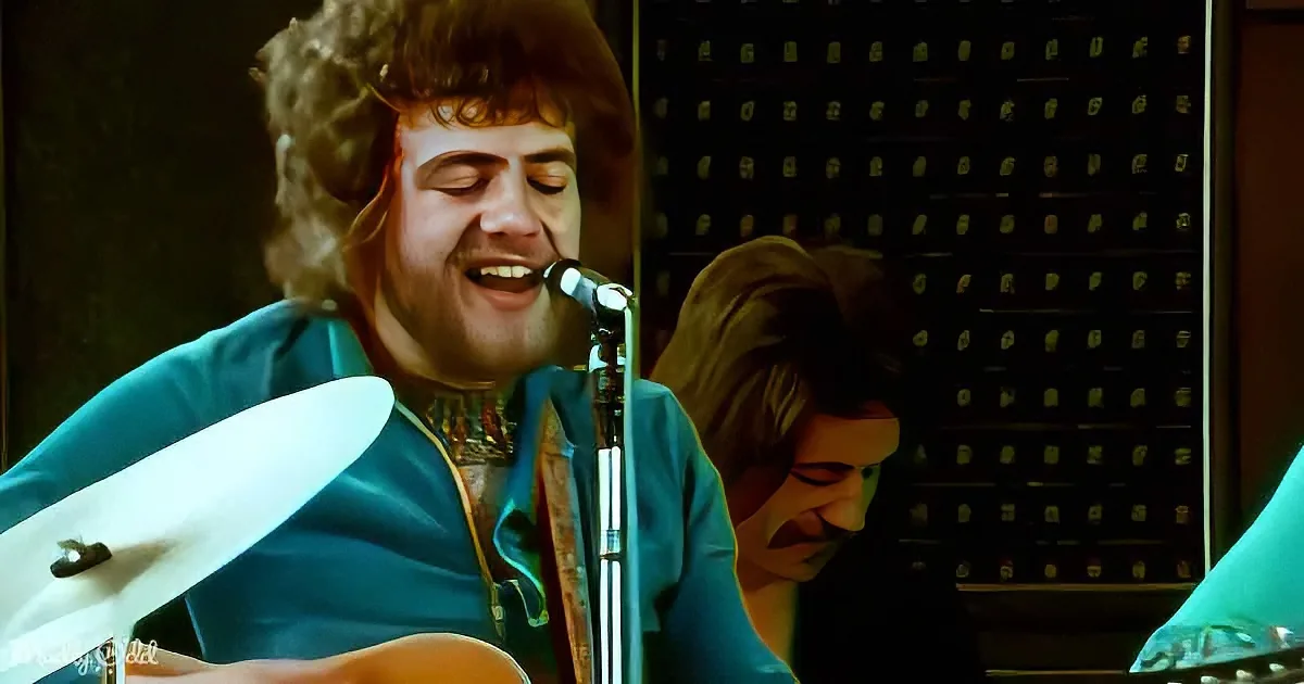 A man in a blue suit plays guitar and sings into a microphone, with another person in the background.