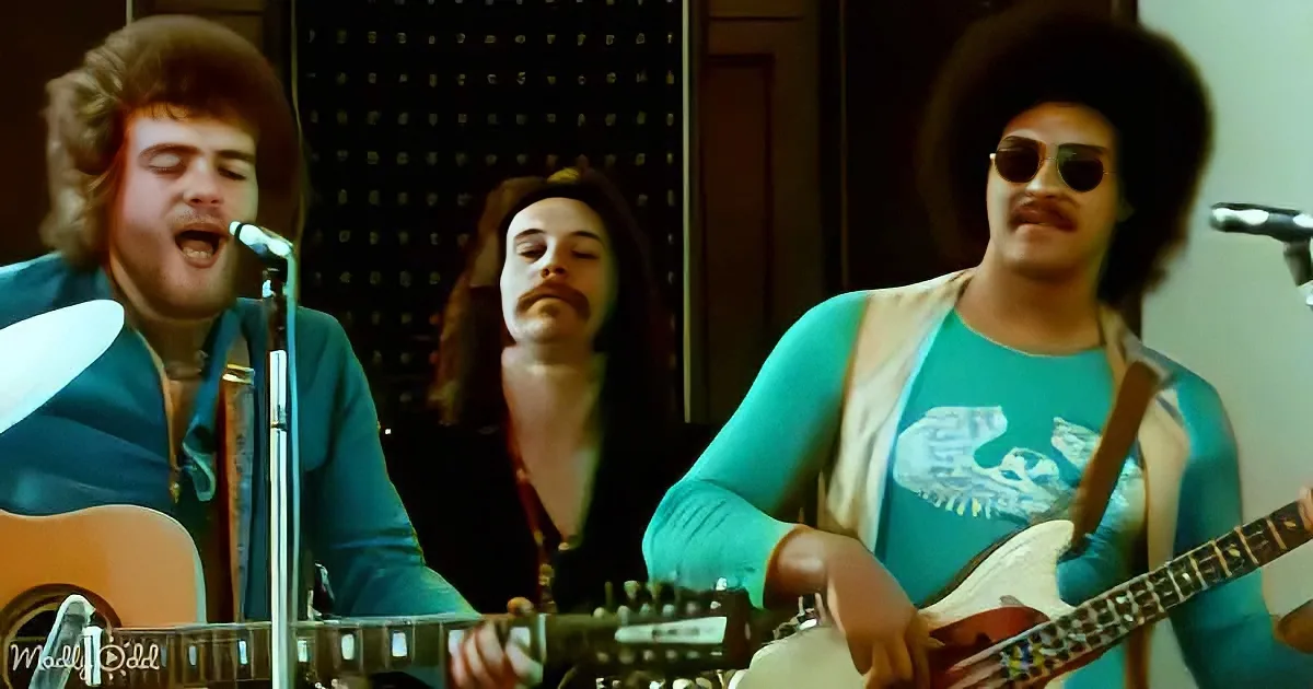 A man in a teal shirt plays guitar while singing into a microphone, with another man in a blue shirt singing behind him.