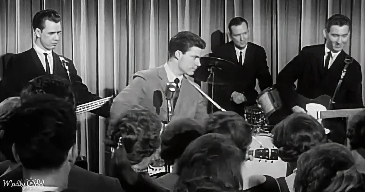 A man in a suit plays guitar on stage, surrounded by a band and an audience.
