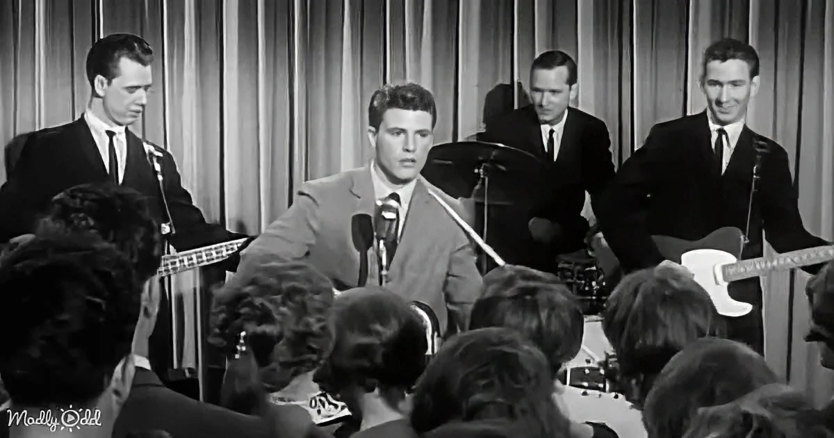 A man in a suit sings into a microphone on stage, surrounded by musicians and an audience.
