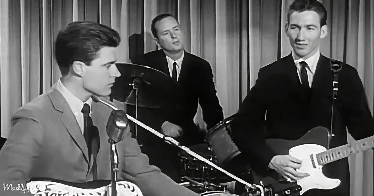A man in a suit sings into a microphone while another plays guitar, with a drummer in the background.