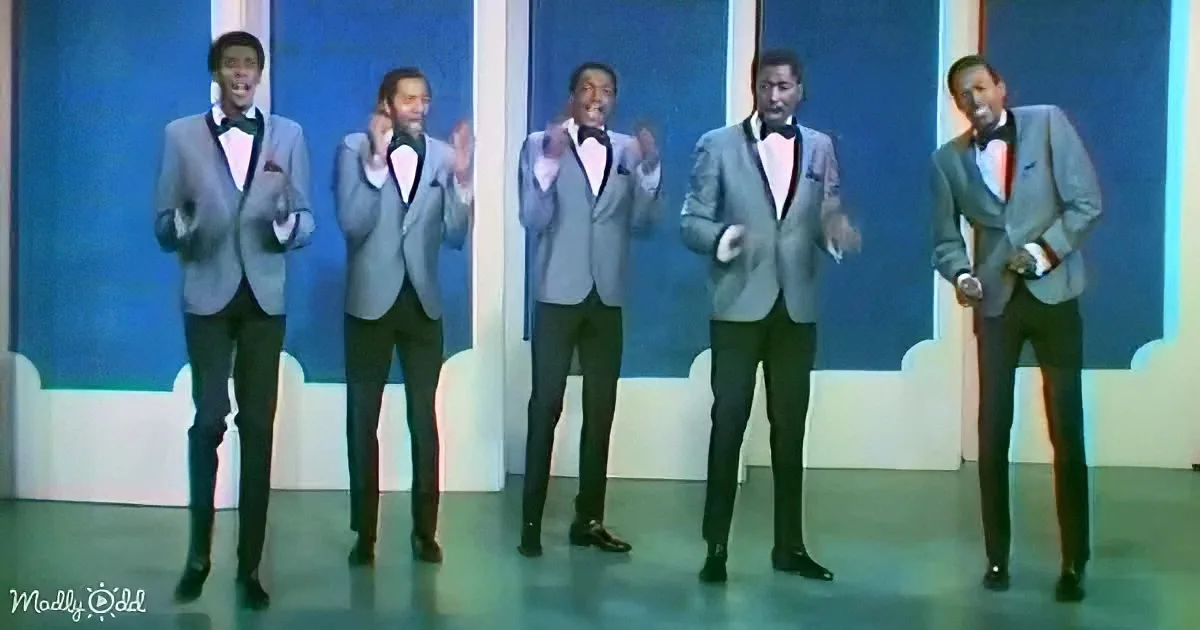 A group of five men in matching gray suits and bow ties perform on stage with a blue and white backdrop.