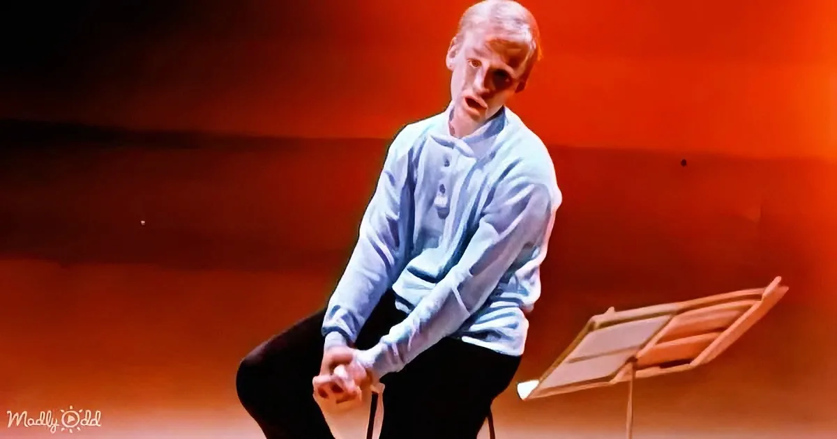A person in a light blue shirt sits on a stool, hands clasped, in front of a music stand, with a red-orange background.