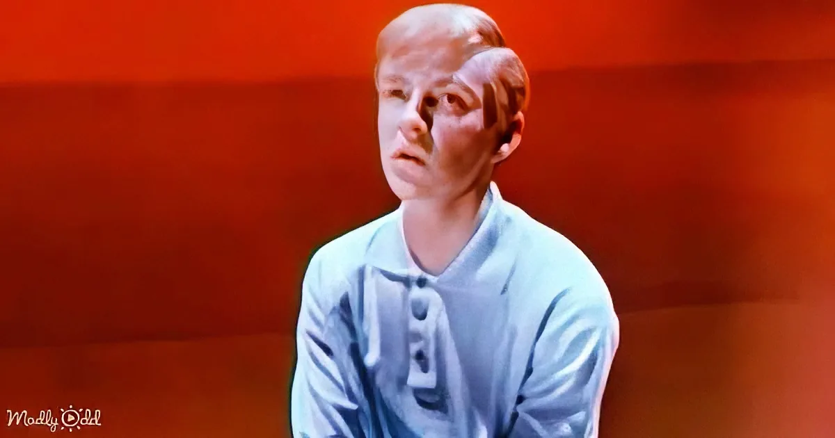 A person in a light blue shirt sits against a red background, appearing to be in a contemplative or thoughtful pose.