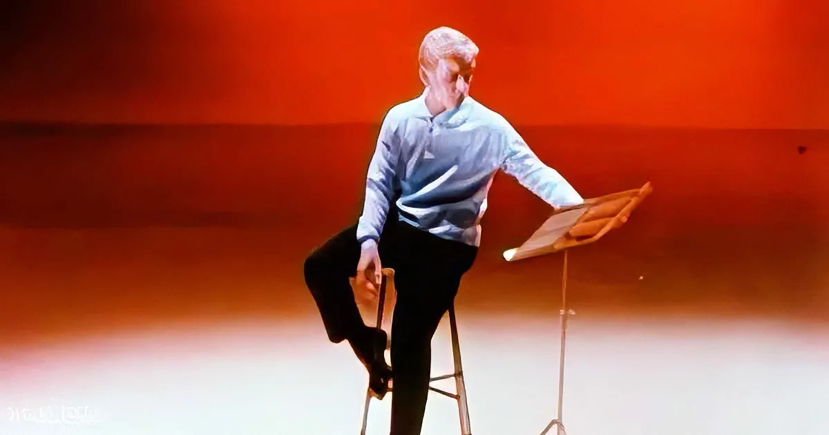 A person in a light blue shirt and dark pants is balancing on one leg on a stool, with a music stand nearby.