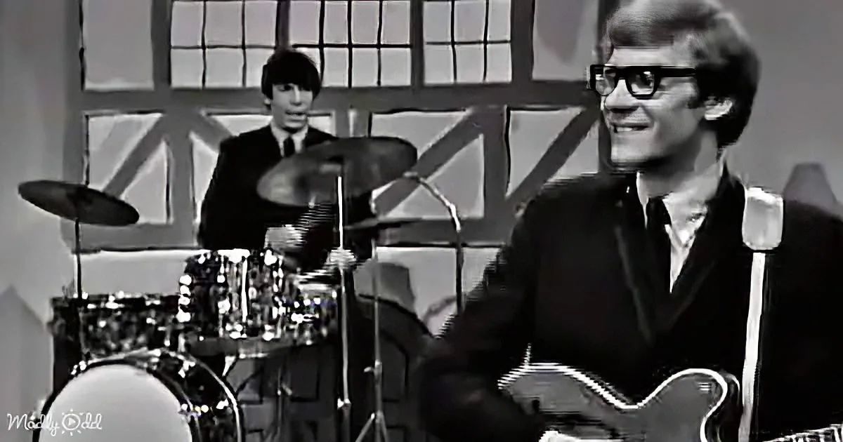 A smiling man in glasses plays guitar, with a drummer behind him in a black suit.