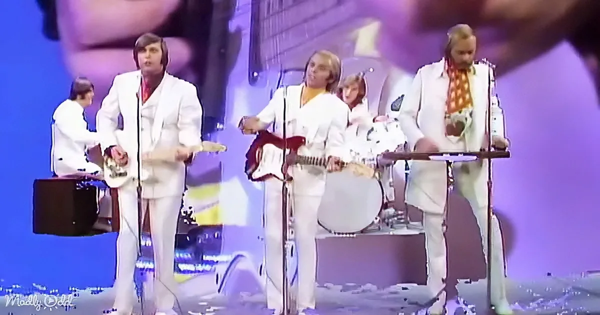 A band in white suits performs on stage with guitars and drums, illuminated by purple and blue lights.
