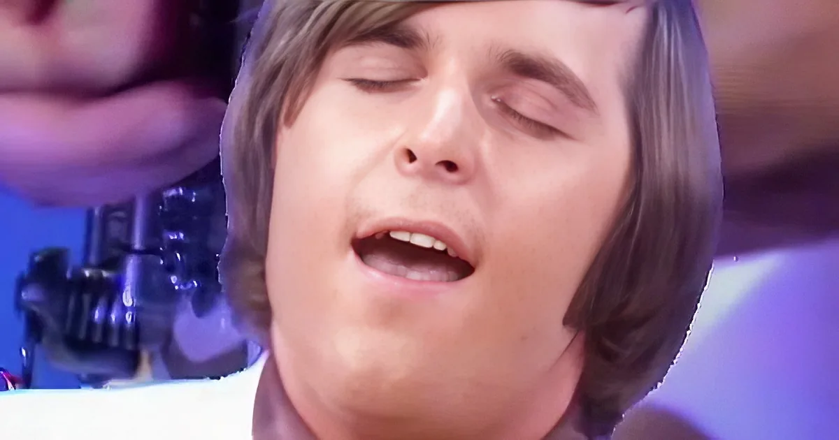 A person with shoulder-length hair and a white shirt is singing with eyes closed, microphone in front.