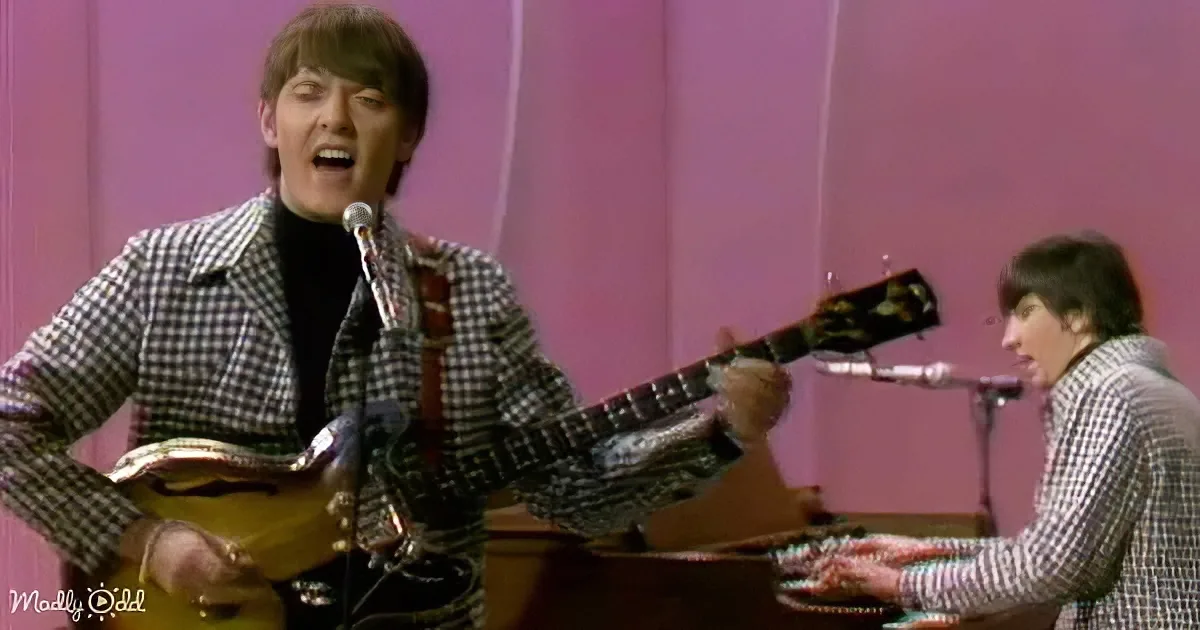 A man in a checkered suit sings into a microphone while playing a guitar.