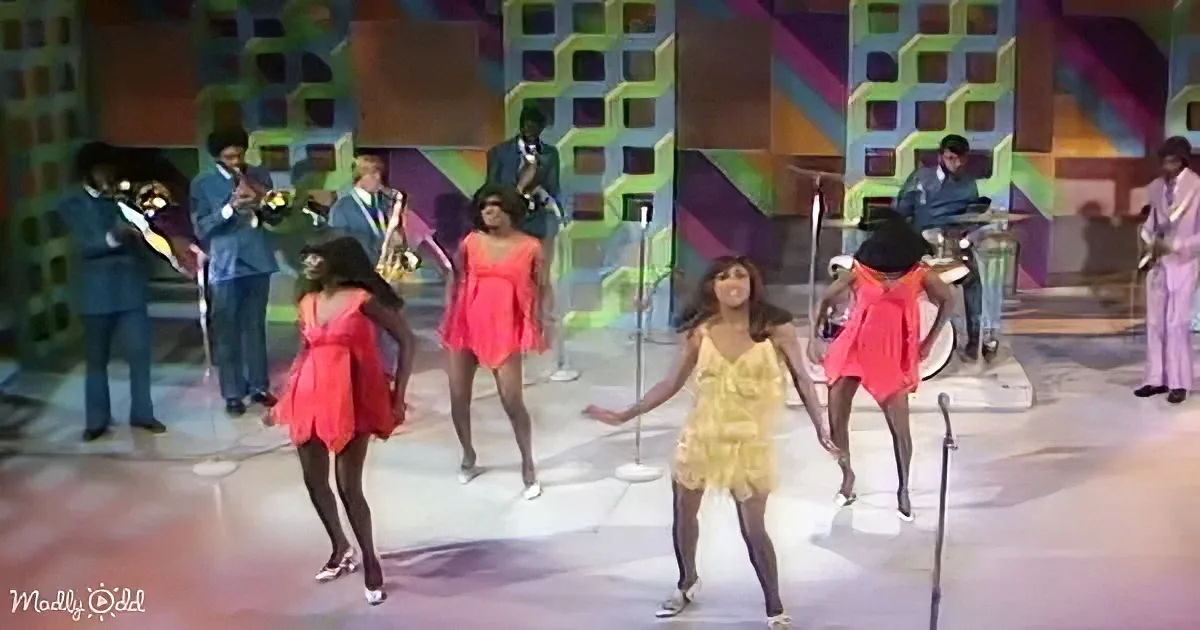 Four women in colorful dresses dance on stage with a band in a vibrant, retro setting.